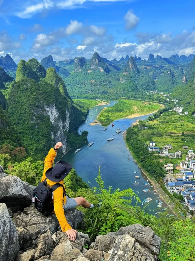 A person wearing a yellow long-sleeved shirt, gray shorts, and a black hat sits on a rocky outcrop overlooking a winding blue river surrounded by lush green karst mountains under a cloudy blue sky. The person is looking out at the landscape with their right arm raised.