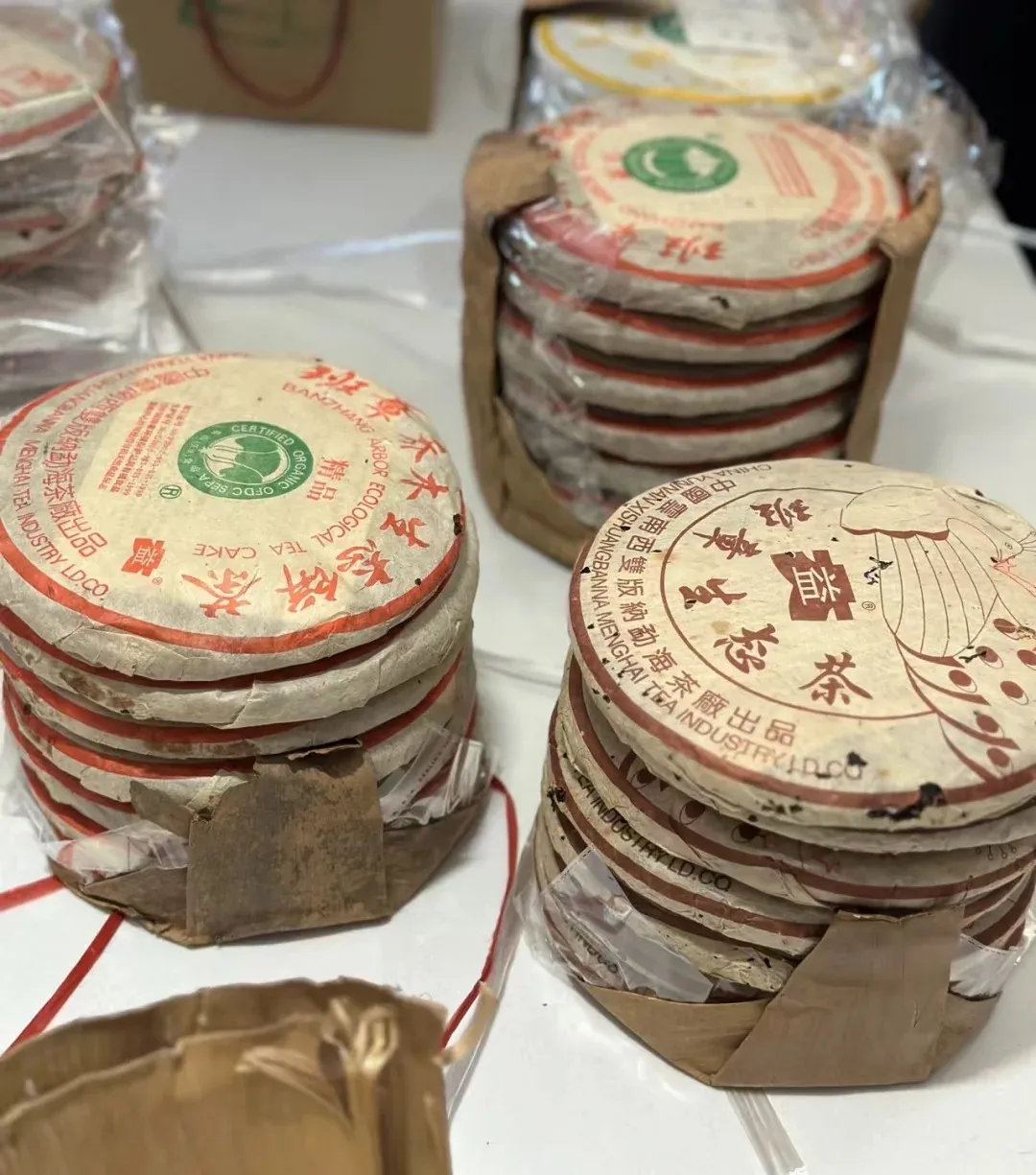 Several stacks of compressed Pu'er tea cakes with red and white circular wrappers. Some wrappers have green certification logos and Chinese characters. The cakes are partially wrapped in brown paper and plastic.