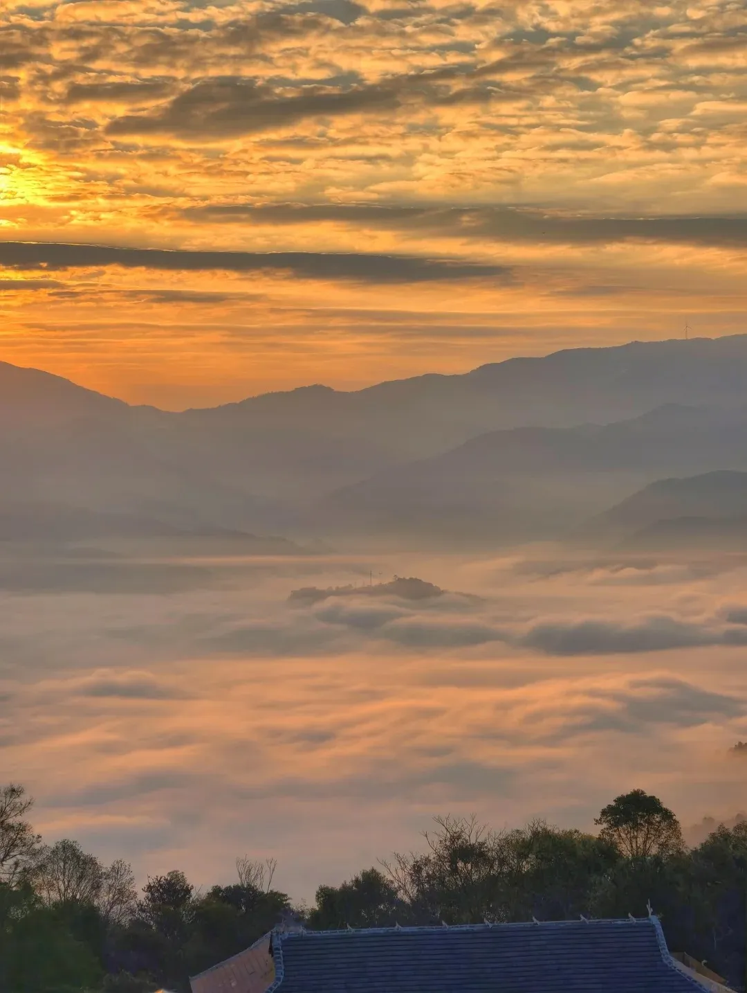 A panoramic view of a valley filled with thick, soft clouds at sunrise. The clouds are a warm orange and pink hue, reflecting the dawn sky. Distant, layered mountains are visible through the mist, fading into the horizon. In the foreground, silhouetted trees and the roof of a building are visible.