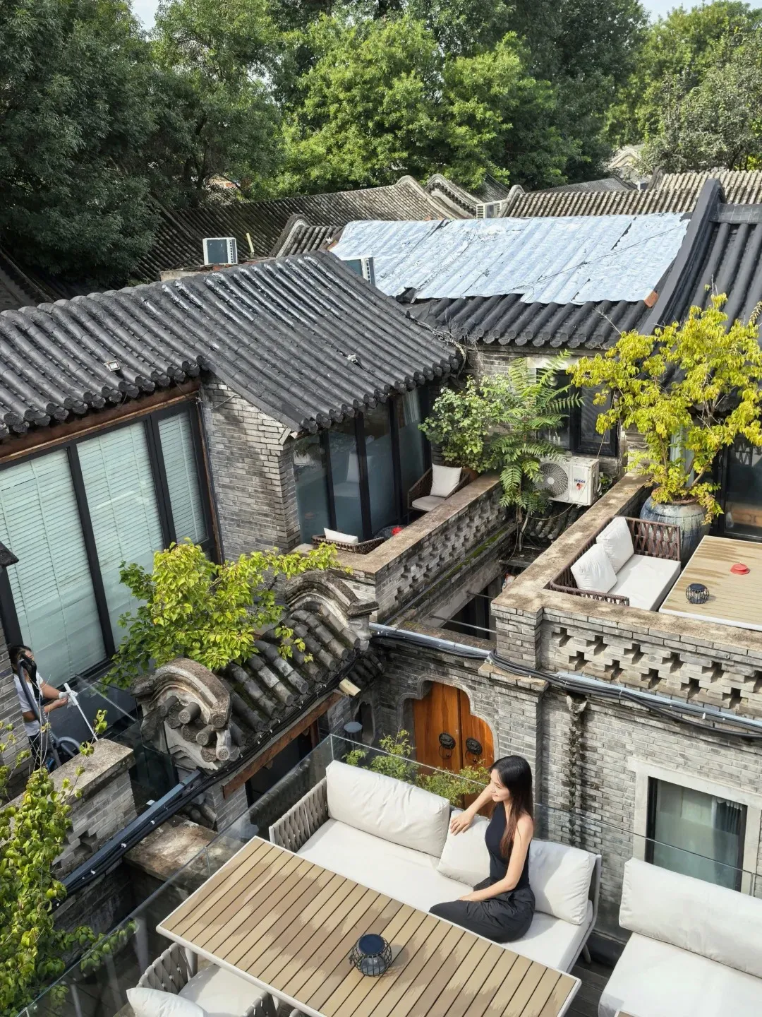 A woman in a black dress sits on a white outdoor sofa on a rooftop terrace overlooking traditional Beijing courtyard rooftops. A wooden table is in front of her, and a person is visible on a lower level cleaning with a vacuum.