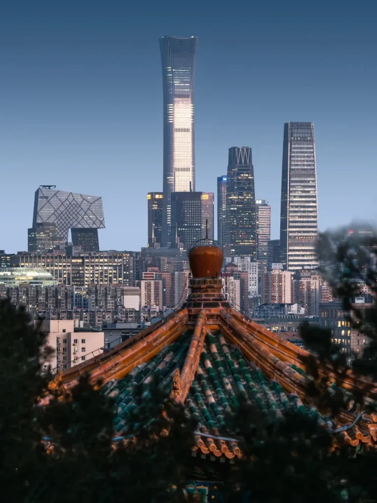 A nighttime view of the Beijing skyline featuring modern skyscrapers, including the China Zun and the CCTV Headquarters, contrasted with the foreground of a traditional Chinese temple roof with green and orange tiles and a golden finial.