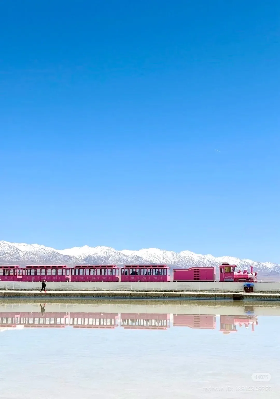 A pink tourist train with multiple carriages travels along a track on a white salt flat. A single person walks near the train. Behind the salt flat is a range of snow-capped mountains under a clear blue sky. The train and mountains are reflected in a still body of water in the foreground.
