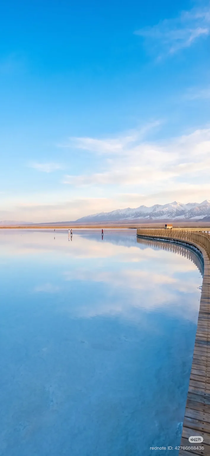 A wide, calm salt lake reflecting a bright blue sky with wispy clouds. In the distance, snow-capped mountains meet a dusty horizon. A curved wooden walkway extends along the right side of the lake. Several figures are walking on the shallow water, their reflections visible.