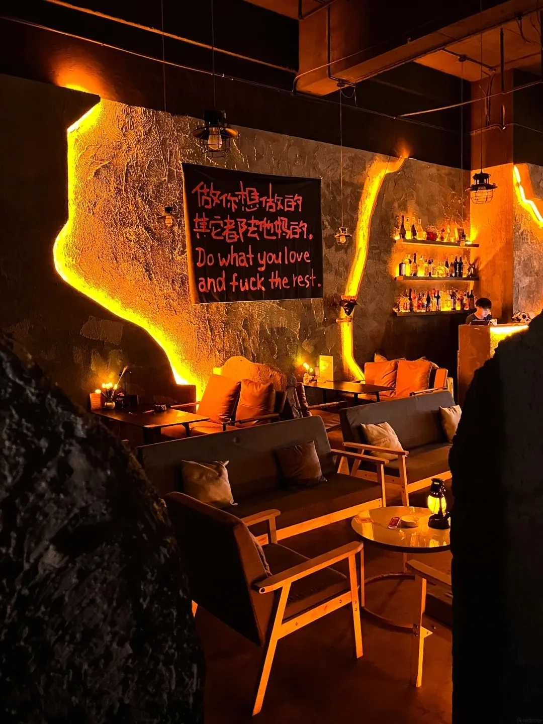 A dimly lit bar interior with textured walls illuminated by bright orange neon lighting. A black banner with red Chinese characters and English text that reads 'Do what you love and fuck the rest' hangs on the wall. Seating includes dark sofas and wooden chairs with cushions, arranged around low tables. Shelves stocked with bottles are visible in the background, along with a person standing behind a counter.