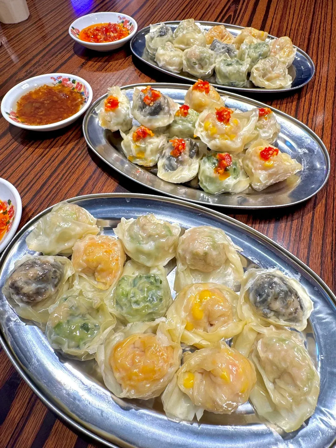 Three metal plates filled with various steamed dumplings, some topped with chili peppers, sit on a wooden table alongside small white bowls of dipping sauce.