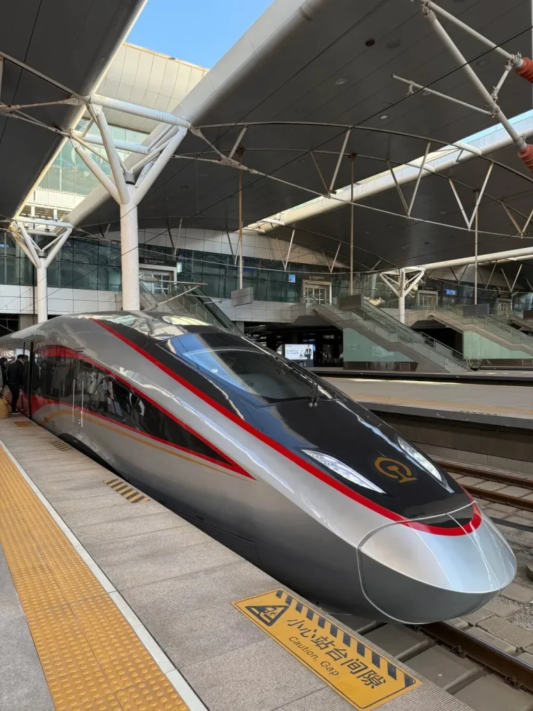 Exterior front view of a silver and red Fuxinghao high-speed train stopped at a station platform under a modern gray and white canopy. A yellow sign on the platform reads 'Caution, Gap'. People are visible near the train doors.