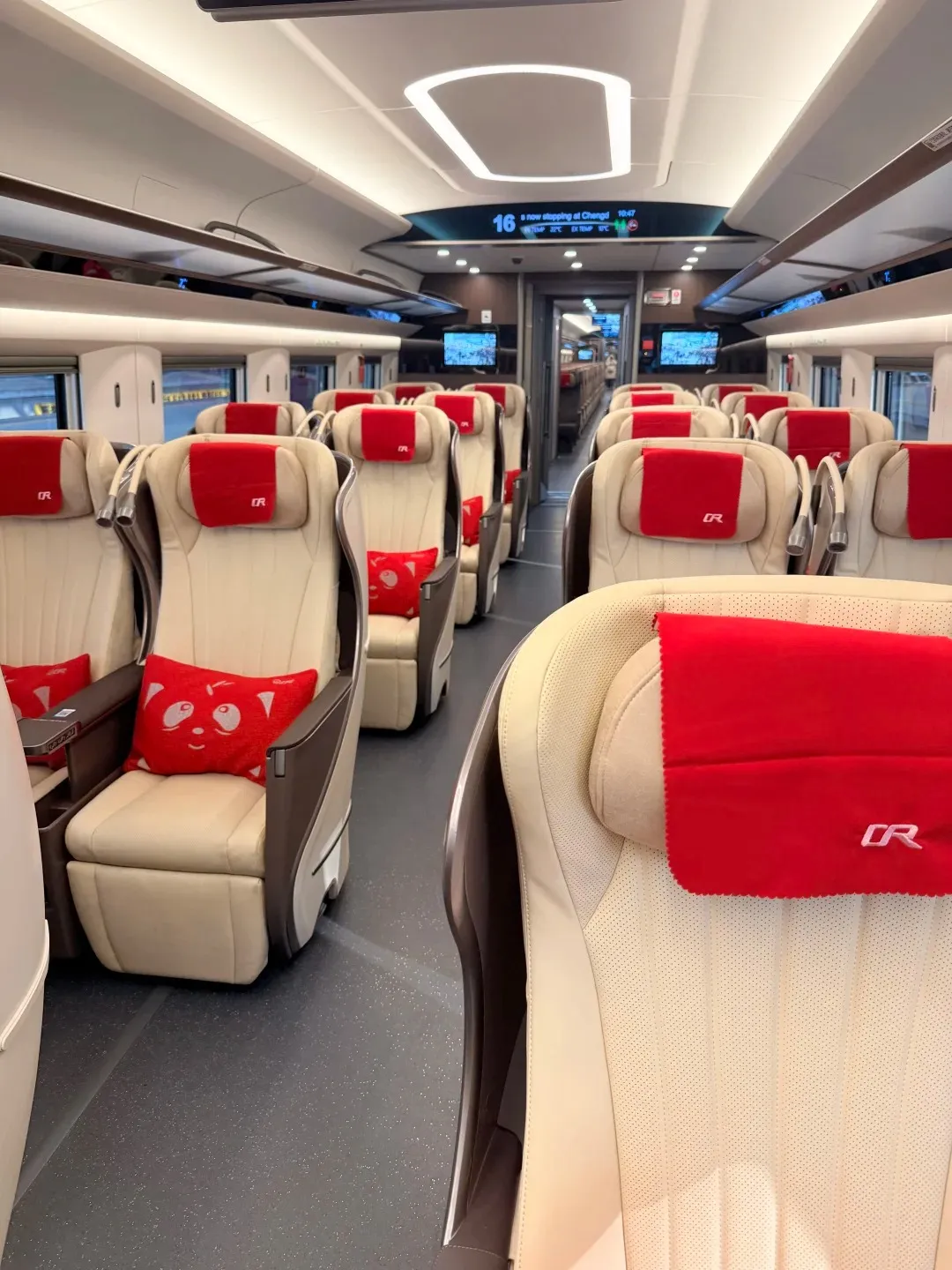 Interior view of a modern high-speed train's silent carriage, showing rows of cream-colored leather seats with red headrests. A few red throw pillows with panda faces are visible on some seats. The aisle is dark gray, and the ceiling features white, illuminated rectangular panels. Digital screens showing train information are visible in the distance.