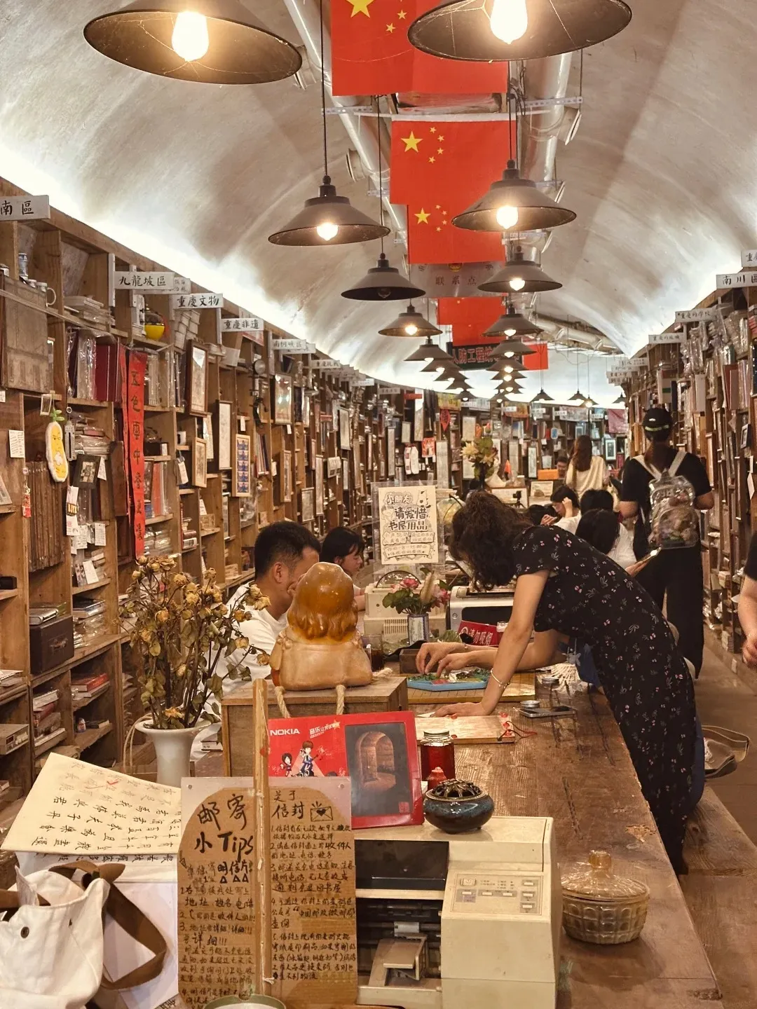 An interior view of a long, arched bookstore built into a converted air-raid shelter. Shelves packed with books line both walls, and Chinese flags hang from the ceiling alongside industrial pendant lamps. A wooden counter in the foreground has various items on it, including a vintage printer, a decorative vase with dried flowers, and signs with Chinese calligraphy. Several people are visible browsing the books and interacting at the counter.