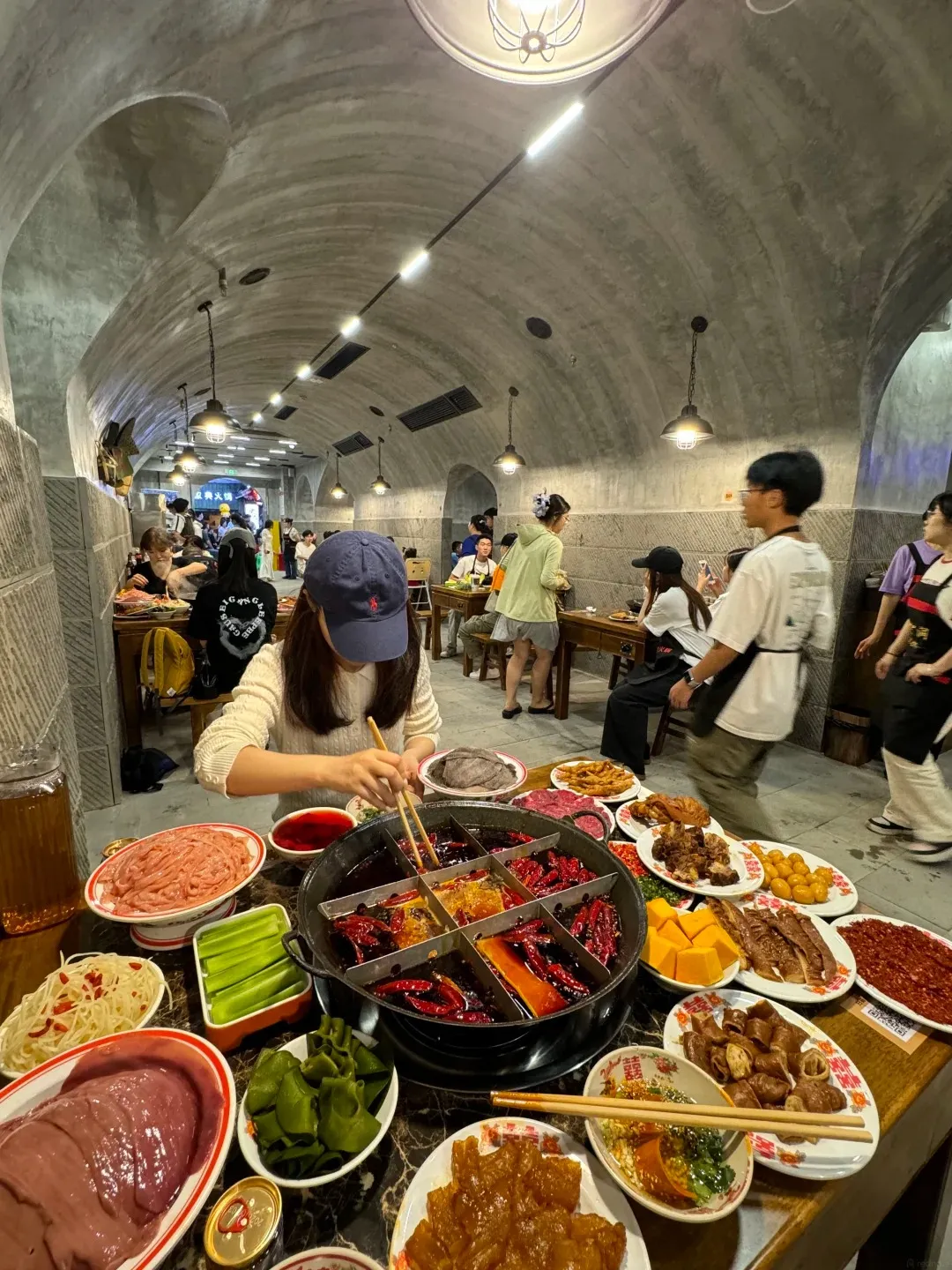 A group of people are dining at a table laden with food, including a large, divided hot pot filled with red broth and various ingredients. Ingredients include sliced raw meat, vegetables like cucumbers and seaweed, and a small dish of dipping sauce. The dining area is inside a large, arched concrete space with visible pipes and industrial lighting, suggesting it is a converted structure.