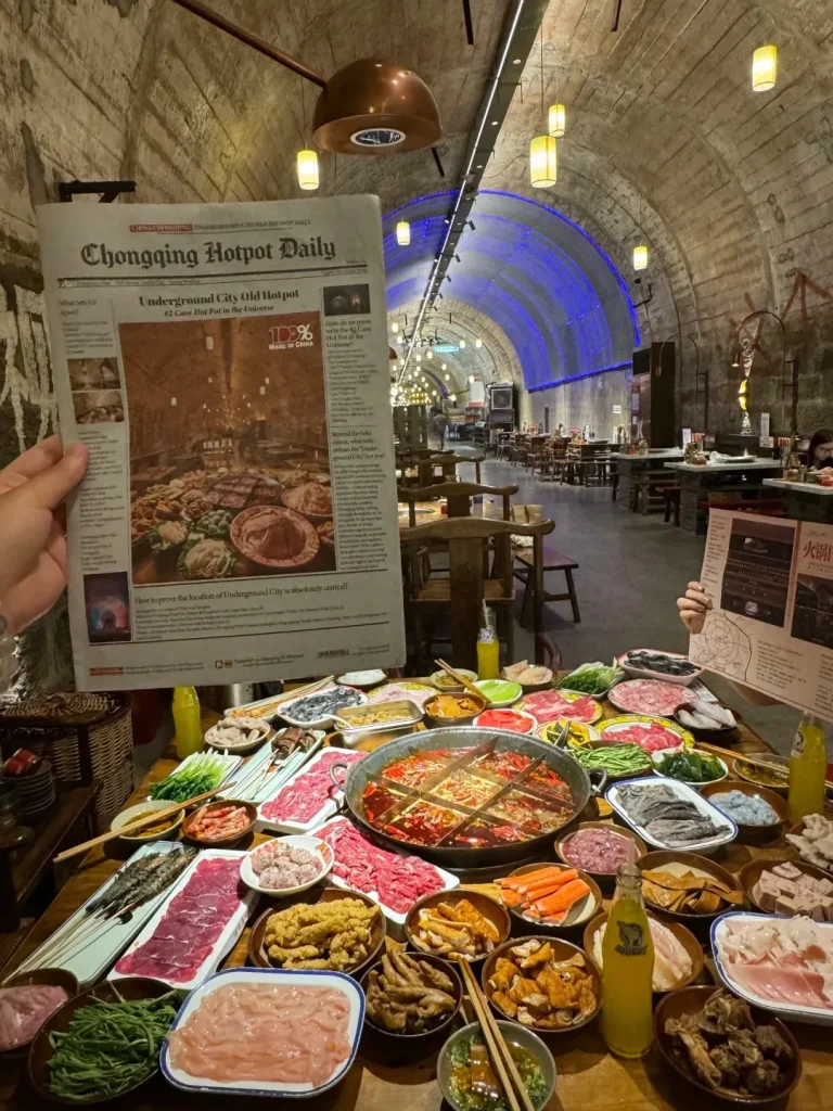 A person holds up a newspaper titled 'Chongqing Hotpot Daily' which features a picture of a large spread of hot pot ingredients. The newspaper is in front of a long table laden with various dishes of raw meats, vegetables, seafood, and other hot pot ingredients, arranged around a central divided hot pot. The table is set in a long, arched concrete tunnel with blue and white lights illuminating the background, suggesting it's a converted air raid shelter. Several bottles of yellow liquid and dipping sauces are also visible on the table.