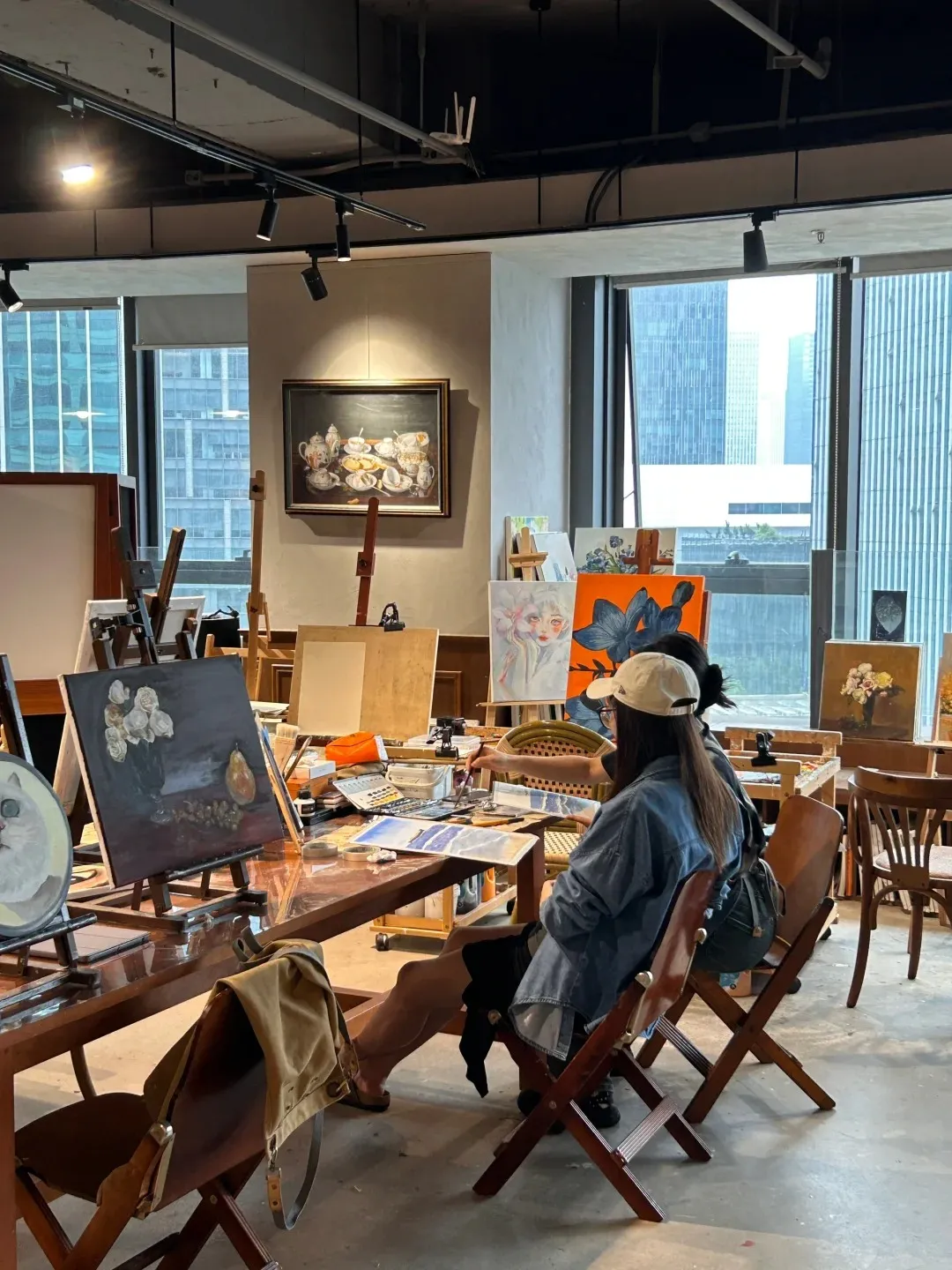 An artist wearing a denim jacket and a baseball cap sits at a wooden table in an art studio, working on a painting. Other easels and paintings are visible in the background, with large windows showing city buildings.