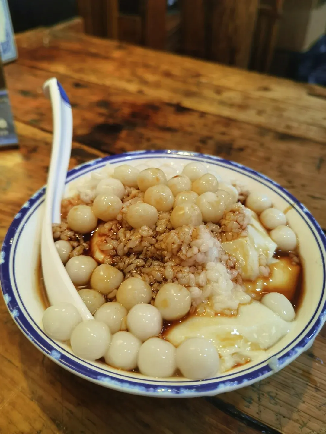 A white bowl with blue trim filled with silken tofu pudding topped with small, round glutinous rice balls, brown sugar syrup, and some cooked rice grains. A white spoon with blue trim rests on the left side of the bowl.