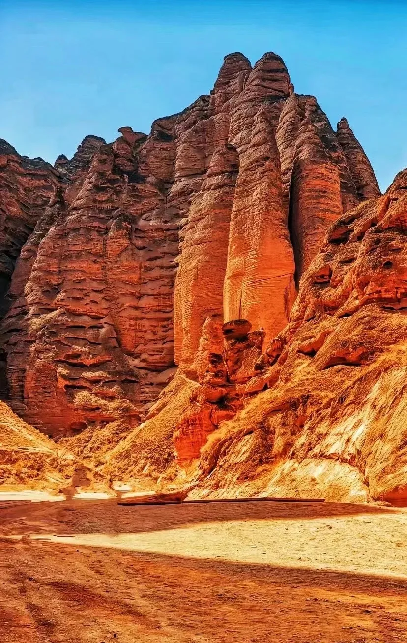 A narrow canyon with towering, striated orange rock formations on either side. The rocks are sculpted by erosion, with many horizontal ledges and overhangs. The canyon floor is sandy and relatively flat, with some shadows cast by the rock formations.