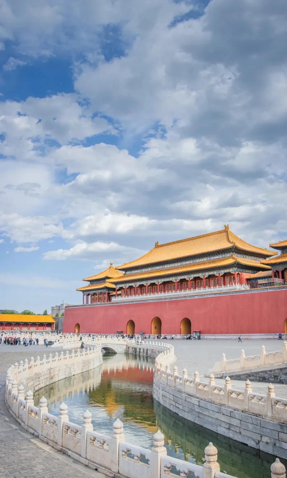 A wide shot of a curved, ornate white stone moat surrounding the red walls of the Forbidden City in Beijing. Golden-roofed traditional Chinese architecture is visible behind the walls, and a white arched bridge crosses the moat. The sky is filled with fluffy white clouds against a blue background.