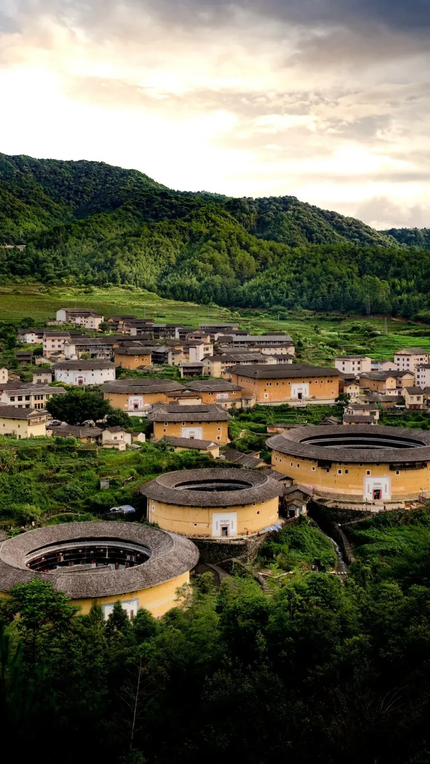 A panoramic view of a traditional Hakka Tulou village nestled in a valley with lush green, terraced hillsides and mountains in the background. Several large, circular, earthen buildings with tiled roofs are visible, along with smaller rectangular houses scattered among them. The foreground shows parts of two large, round Tulous, one more prominent and in focus than the other.