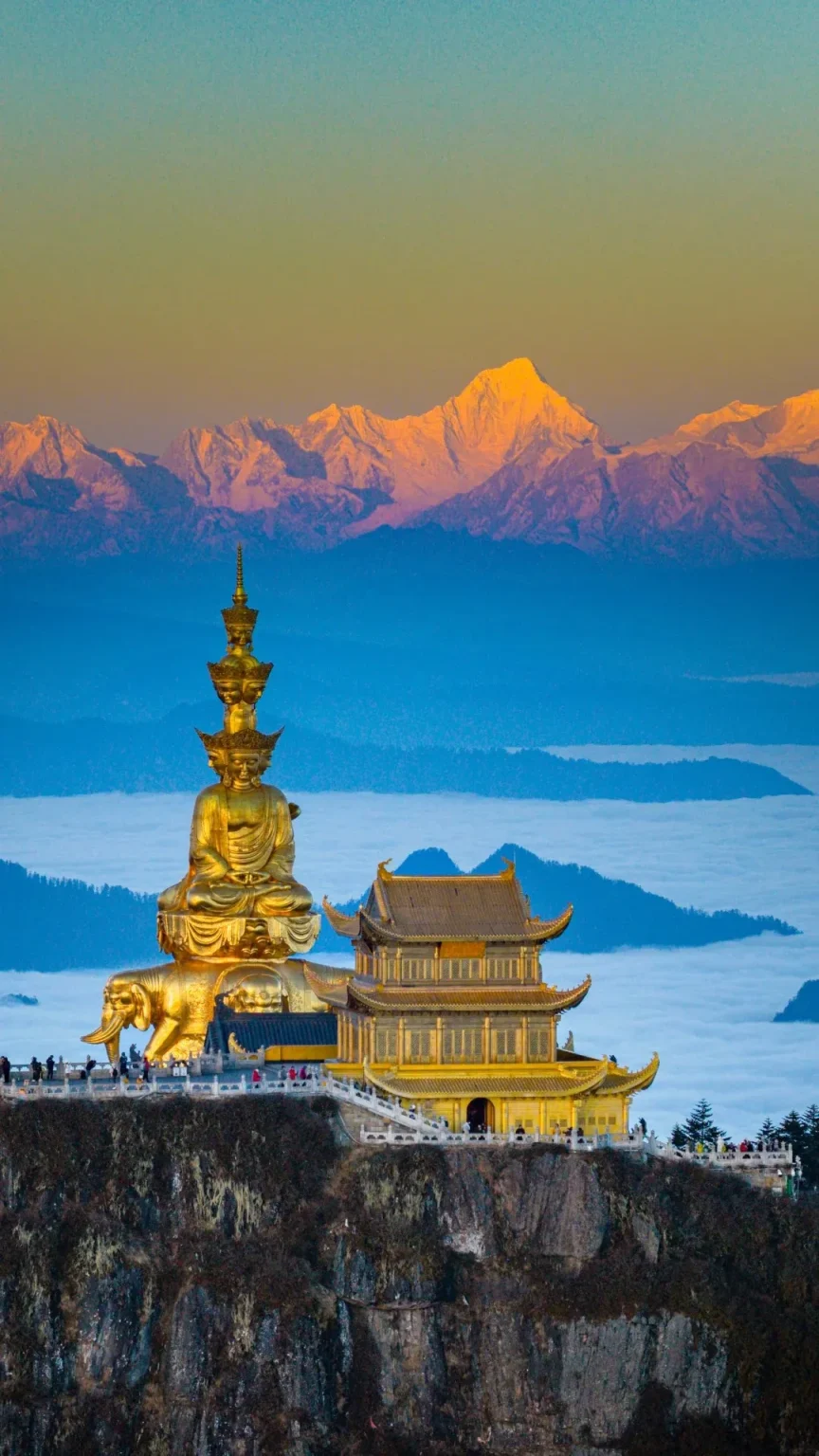 A majestic, multi-tiered golden Buddhist statue of Samantabhadra is seated on an elephant base, located on a rocky mountain peak. To its right stands a golden traditional Chinese temple. In the background, layers of misty clouds fill the valleys, and a breathtaking mountain range is illuminated by the golden light of sunrise.