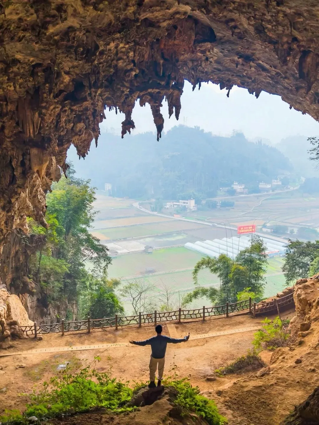View from the opening of a large natural cave, looking out over a misty valley with terraced fields, small villages, greenhouses, and hills in the background. A person stands at the cave entrance with arms outstretched.