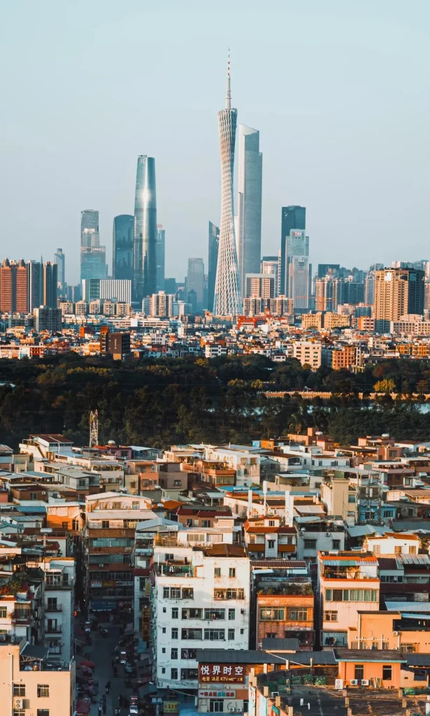 A panoramic aerial view of Guangzhou, China, featuring a dense urban landscape. In the background, several modern skyscrapers dominate the skyline, including the distinctive Canton Tower with its lattice structure. In the foreground and mid-ground, there are numerous tightly packed residential buildings with varied architectural styles and colorful facades, interspersed with trees and some signs of street life.