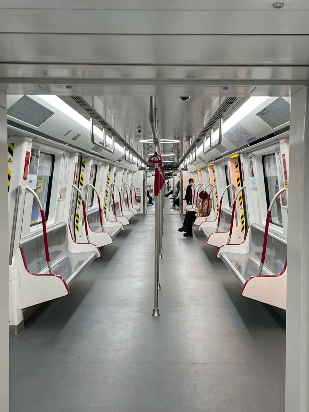 Interior view of a modern, clean Guangzhou Metro train carriage with rows of white and red seats lining both sides. Several passengers are seated, and a few are standing near the doors. The floor is gray, and the ceiling has bright white lighting and silver handles.