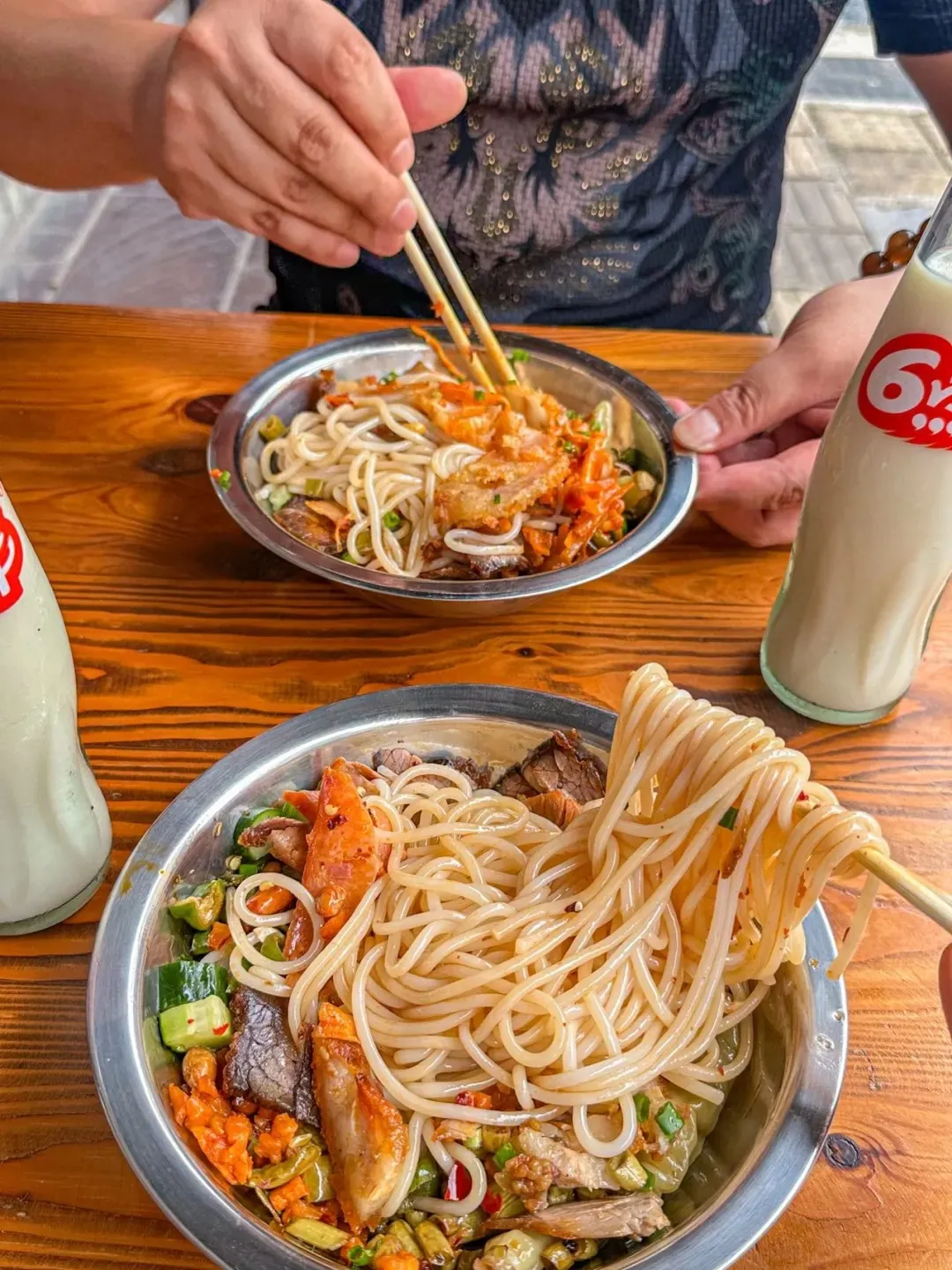 Two stainless steel bowls of Guilin rice noodles mixed with meat and vegetables sit on a wooden table. One bowl has chopsticks lifting noodles. Two glass bottles of a white beverage are on either side of the bowls.