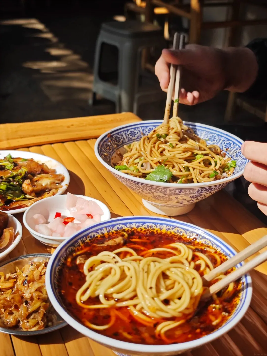 A close-up of two bowls of Guilin rice noodles on a bamboo table. One bowl in the foreground is filled with noodles in a spicy red broth, and is being lifted with chopsticks. A second bowl of noodles with green onions and sauce is behind it. Several small side dishes of pickled vegetables and other toppings are arranged around the bowls.