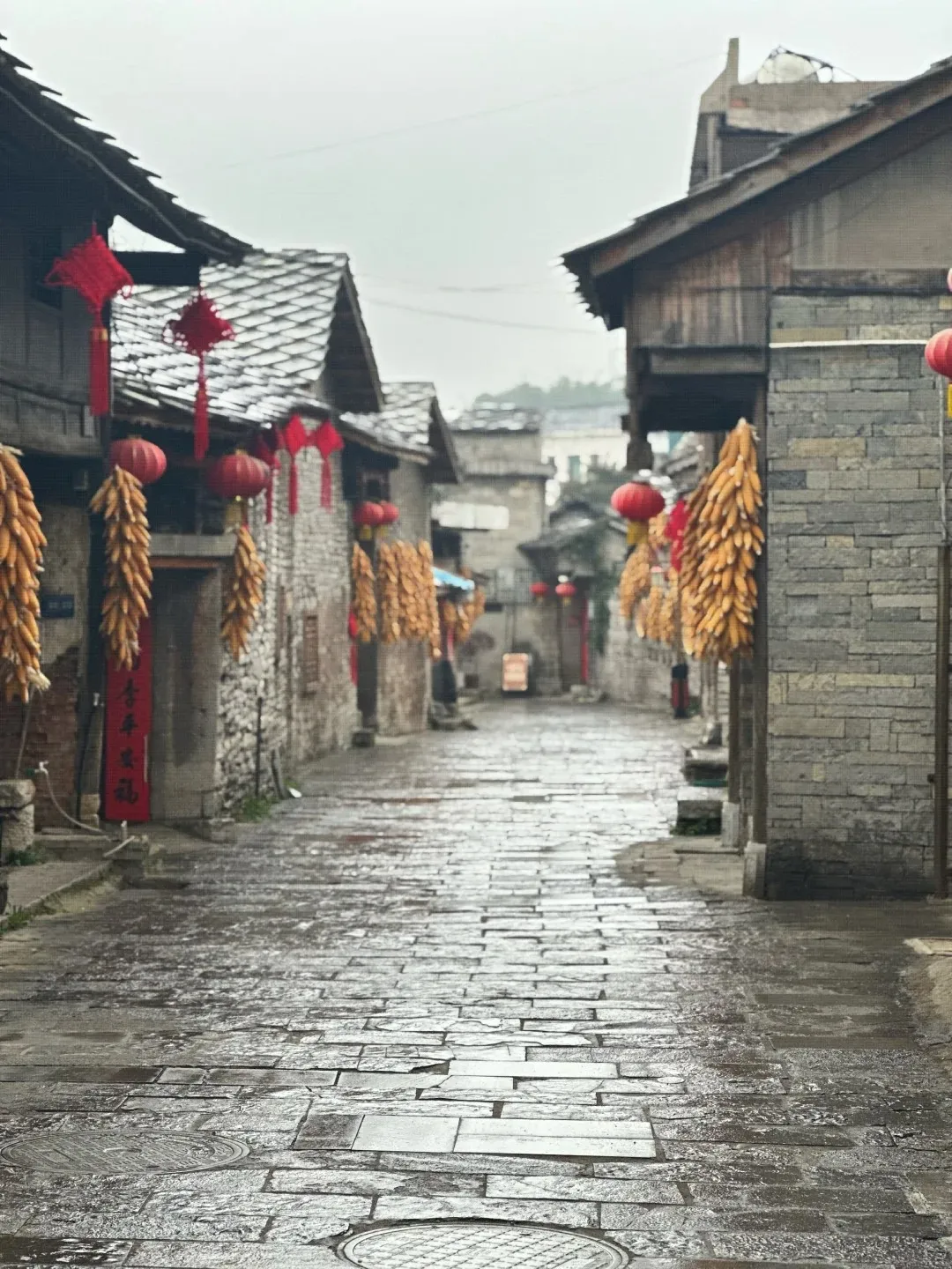 A wet, stone-paved alleyway in an ancient Chinese village, lined with traditional stone and wood buildings. Bunches of dried corn and red lanterns hang from the eaves.