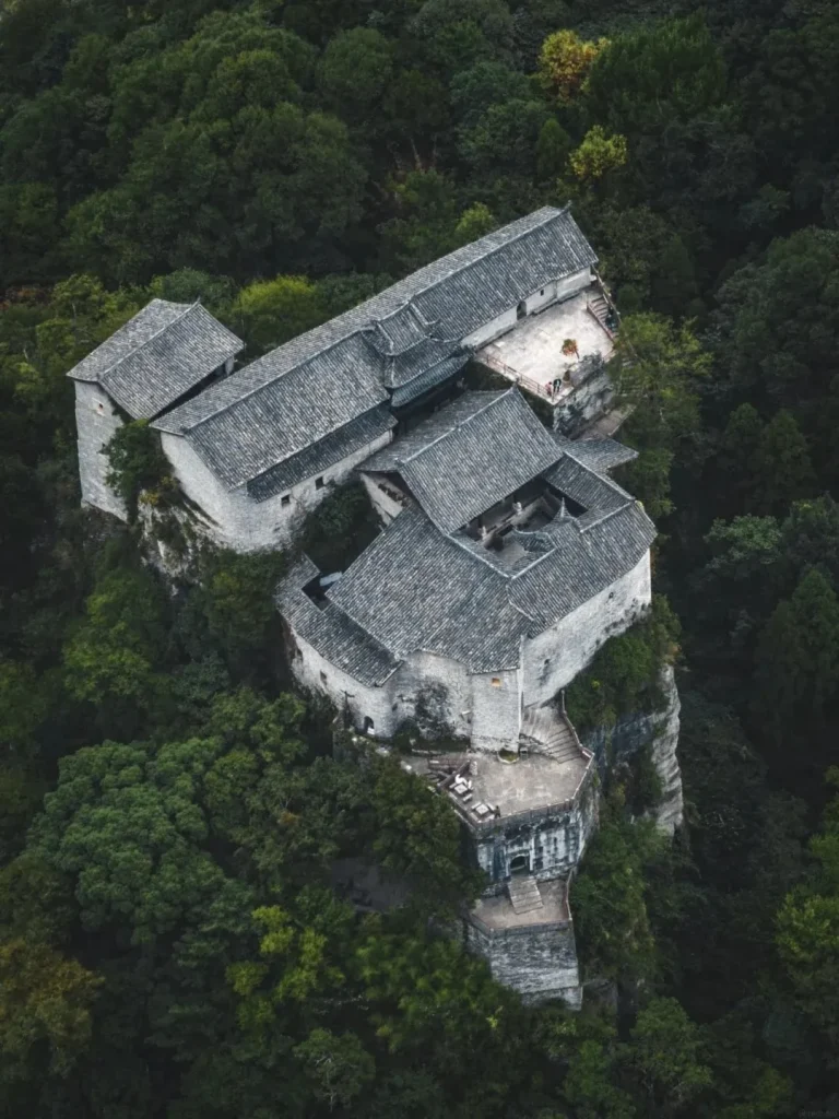 An aerial view of an ancient stone village complex built on the side of a steep, forested mountain. The architecture features grey tiled roofs, stone walls, and courtyards, with narrow staircases and patios clinging to the cliff face. Lush green trees surround the structures.