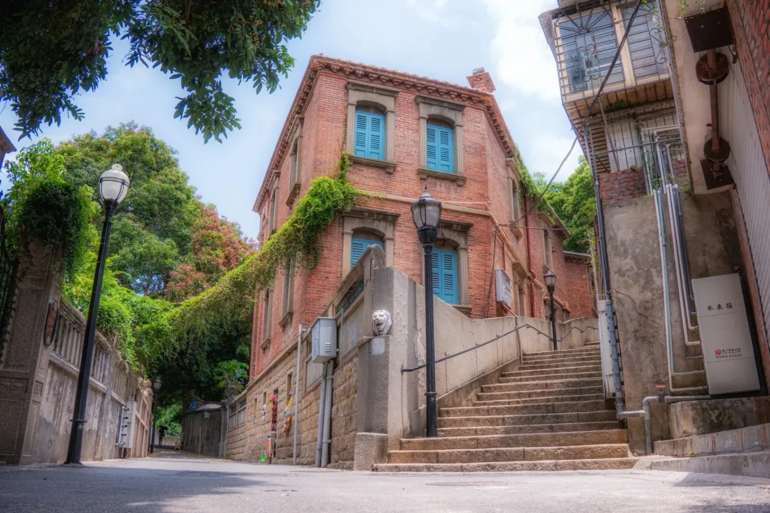 A wide-angle view of a narrow street on Gulangyu Island with a prominent red brick building with blue shutters and ivy climbing its walls. A stone staircase leads up to the building on the right. A lamppost stands to the left of the stairs, and another lamppost is visible further down the street. Lush green trees and foliage surround the buildings and line the street.