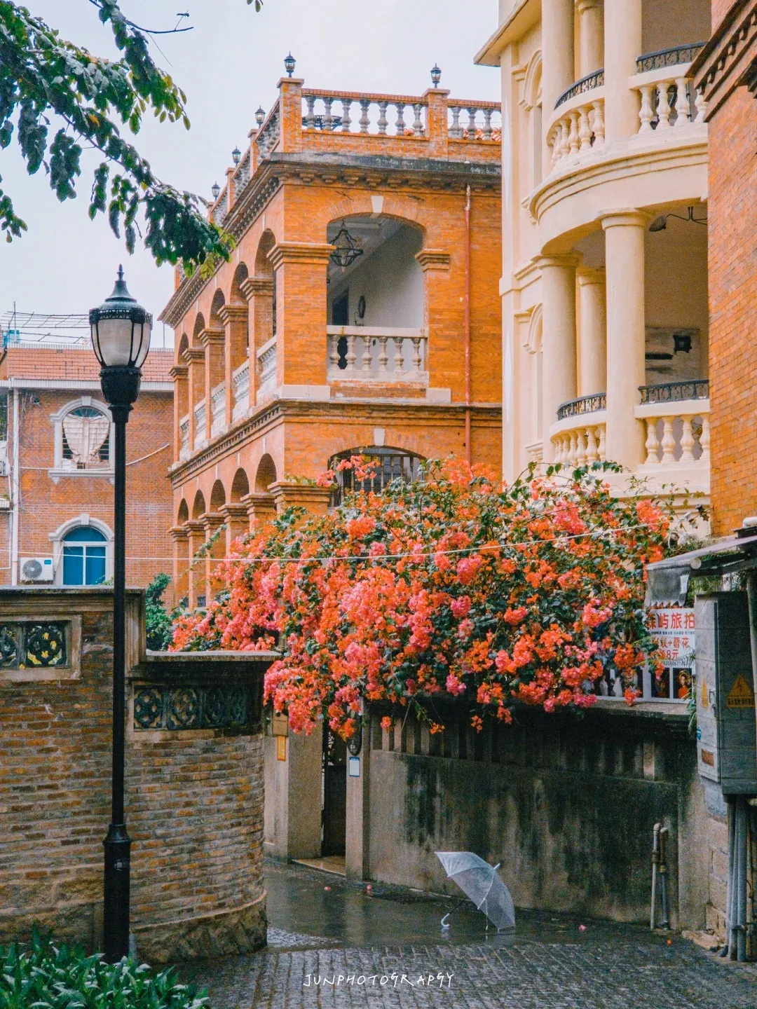 An ornate red brick European-style building with arched windows and balconies stands next to a lighter colored building with classical columns. A large bush of vibrant pink bougainvillea flowers cascades over a low stone wall. A black lamppost is in the foreground, and a transparent umbrella lies open on a wet cobblestone path. The sky is overcast.