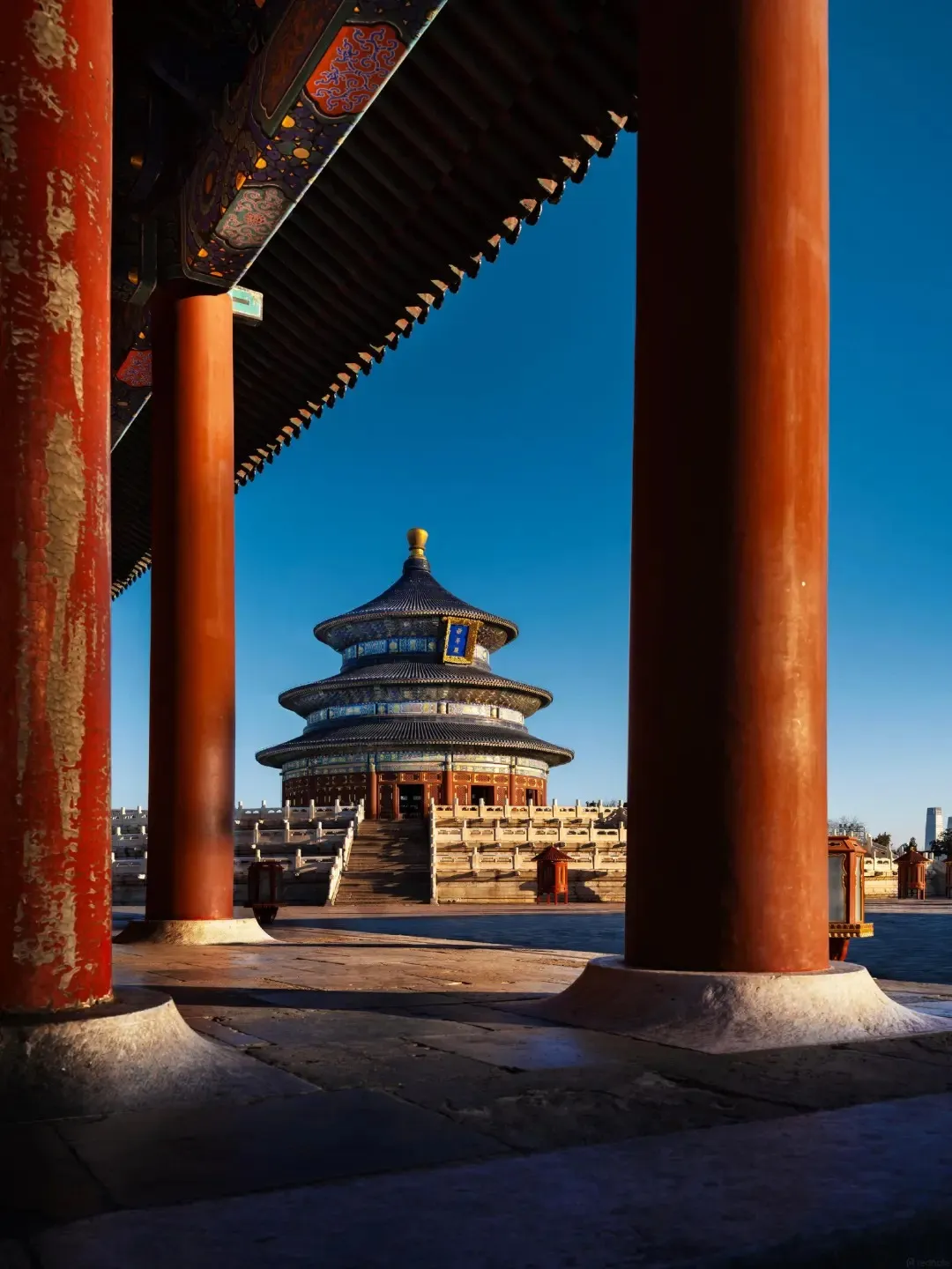 Framed by two large red pillars in the foreground and a shadowed wooden eave overhead, the triple-eaved Hall of Prayer for Good Harvests at the Temple of Heaven in Beijing is visible under a clear blue sky. The circular building has a dark blue tiled roof topped with a golden finial. It sits on a multi-tiered white marble terrace with stone balustrades and stairs leading up to its entrance. Several smaller red lanterns are placed around the terrace.