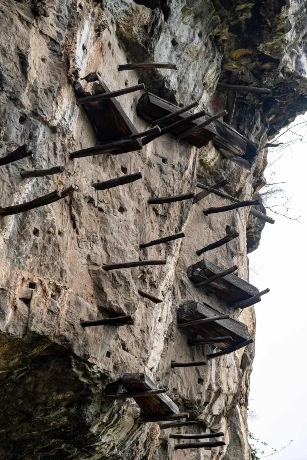 Numerous wooden coffins are precariously balanced on wooden beams projecting from a steep, rocky cliff face.