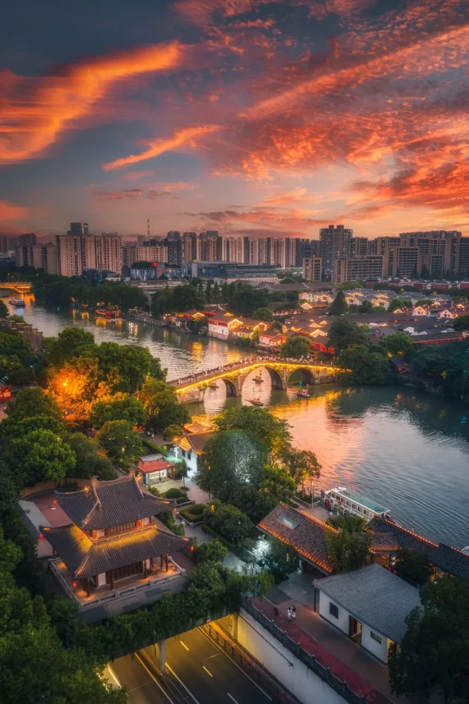 A wide shot aerial view of the Grand Canal in Hangzhou at sunset. A traditional arched bridge, lit by numerous golden lights, spans the canal, with boats passing underneath. The banks of the canal are lined with traditional Chinese buildings with tiled roofs and illuminated windows. Lush green trees surround the buildings and the canal. In the background, a dense cityscape of modern high-rise buildings rises against a vibrant orange and red sky filled with dramatic clouds.