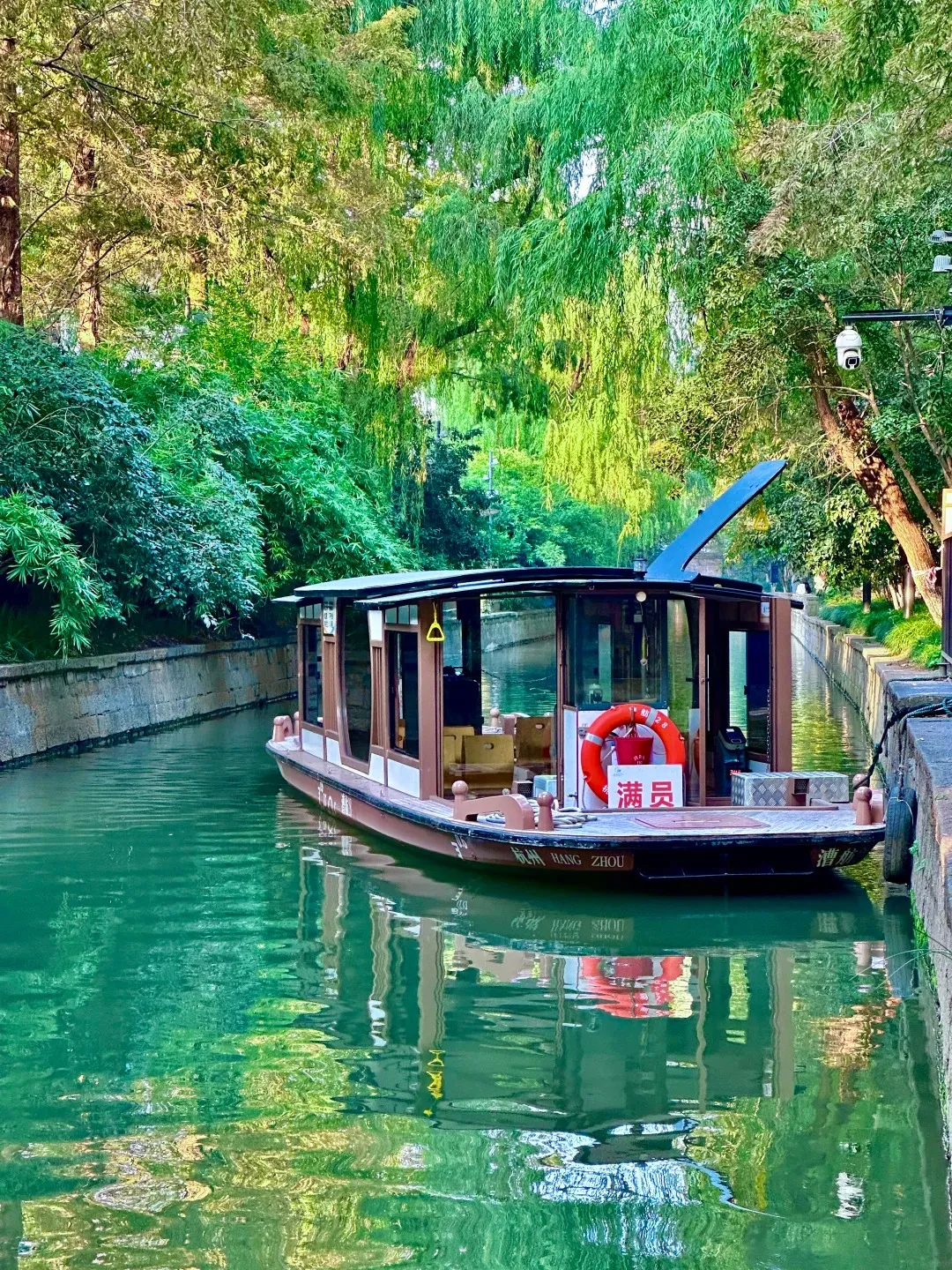 A narrow boat with a brown and white cabin and a dark roof with a blue fin sails on a green canal. The boat has windows and a life preserver hanging on the side. Lush green trees with long, drooping branches line both sides of the canal, and their reflections are visible in the water.