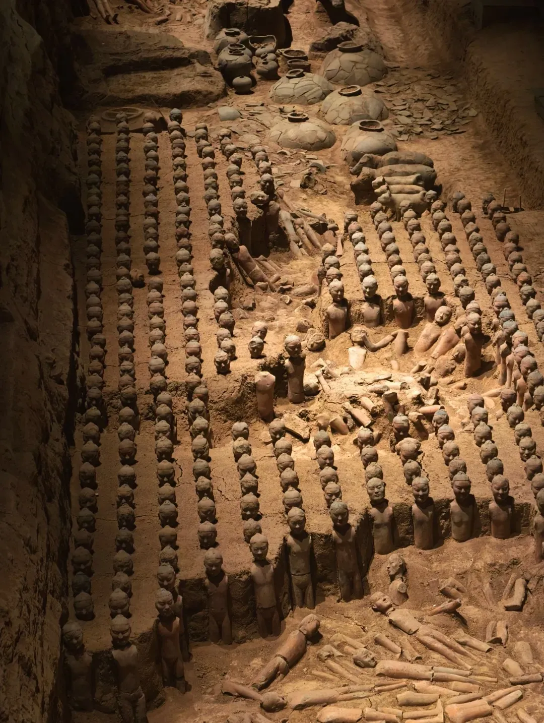 An aerial view of an archaeological excavation pit containing rows of miniature terracotta figures and pottery jars. Some figures are standing in formation, while others are lying down. The pit is made of brown earth, with some areas covered by overhead structures and debris. Sunlight or artificial light illuminates parts of the scene.
