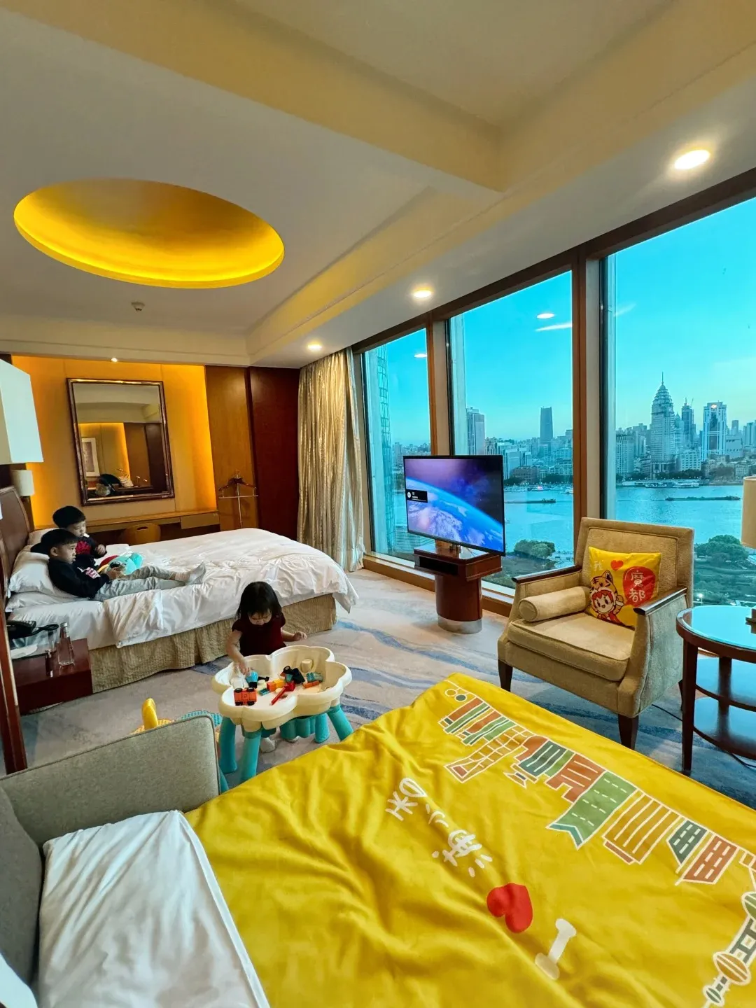 A hotel room with two beds, one with children on it, a child playing with toys on the floor, a television, and a large window overlooking a city skyline and river at dusk.