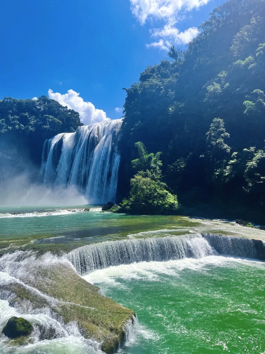 A wide, powerful waterfall cascading over a cliff face, surrounded by dense green trees and a bright blue sky with white clouds. The water falls into a turquoise pool at the bottom, with smaller tiered waterfalls in the foreground.