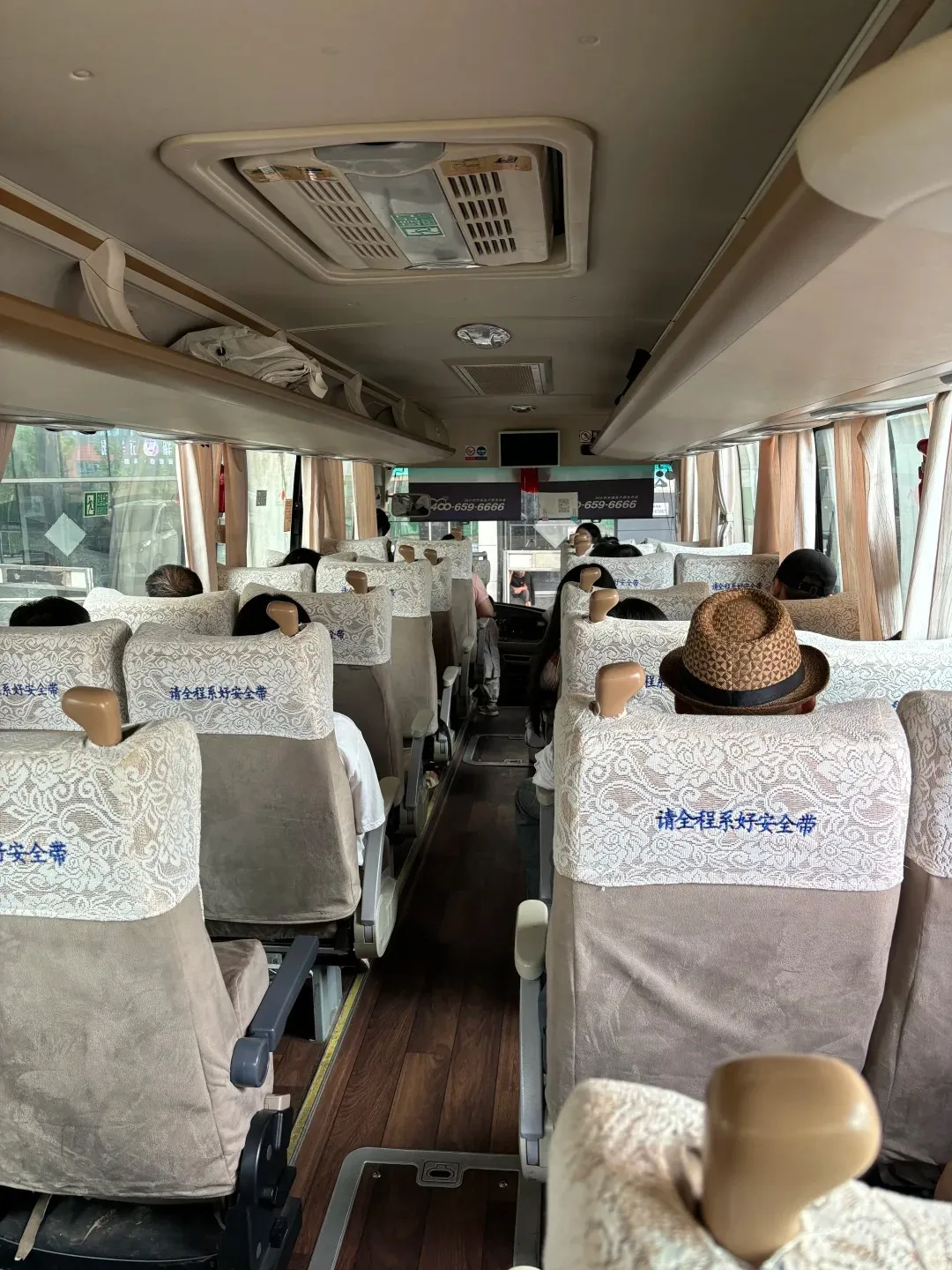 The interior of a bus shows rows of seats with light brown upholstery and white lace-covered headrests. Several passengers are visible, some wearing hats. The aisle has a wooden floor. Overhead compartments and air vents are also visible.
