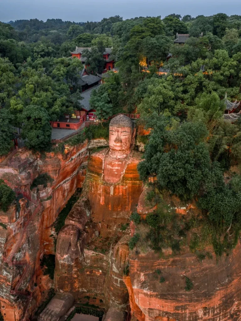 Aerial, wide-angle view of the Leshan Giant Buddha, a massive stone statue carved into a cliff face. The Buddha is seated, facing forward, with intricate details visible on its face and torso. The statue is made of reddish-brown sandstone and is surrounded by lush green trees and traditional red-roofed temple buildings situated on the cliff above and to the side.