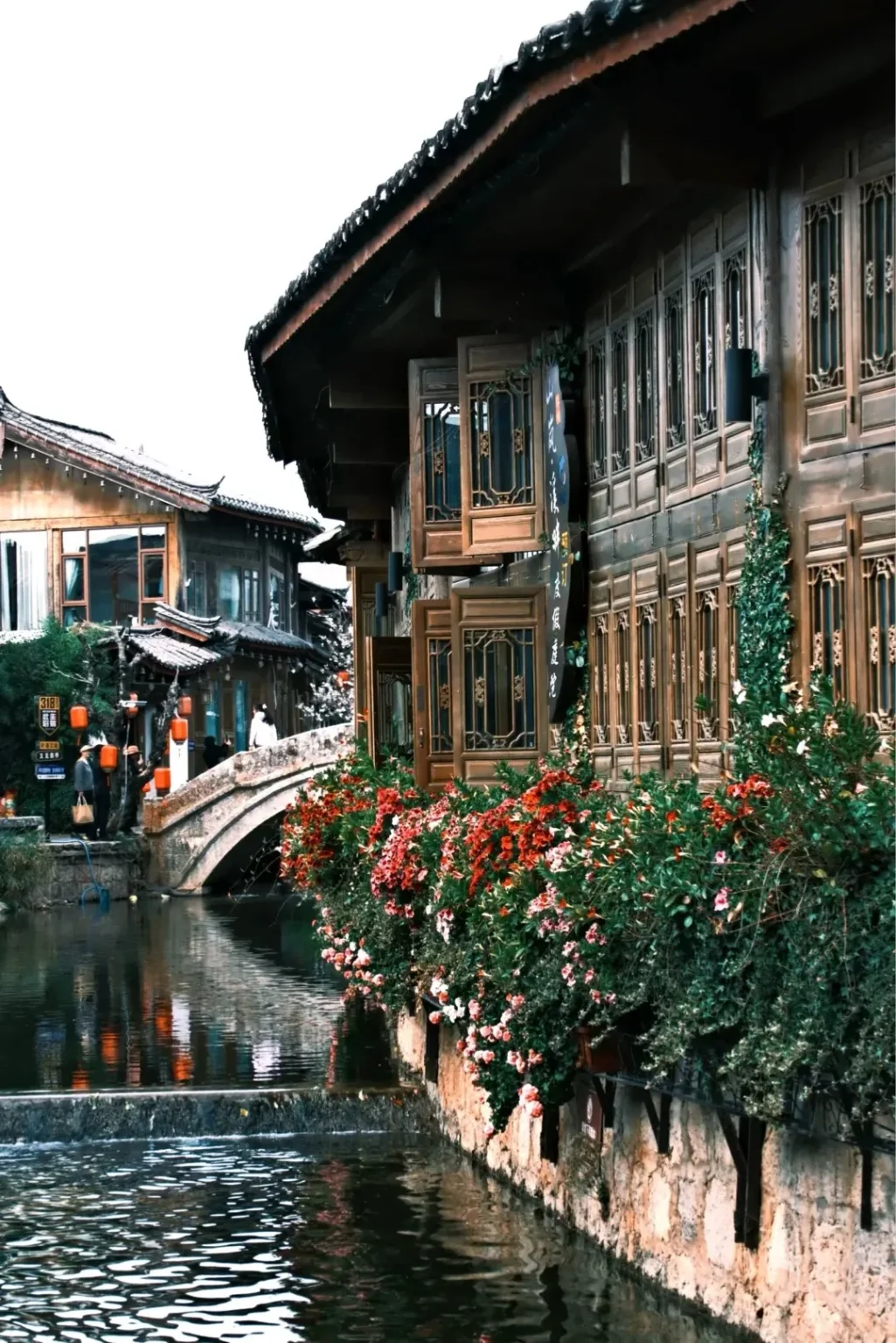 A canal with reflections runs along traditional wooden Chinese architecture. A stone bridge arches over the canal in the background, with a lush display of red and pink flowers cascading from the buildings.