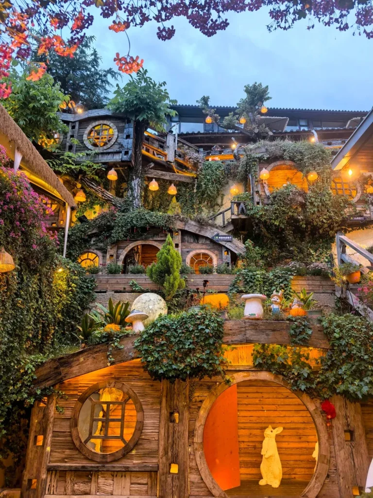 A multi-story building with a whimsical, hobbit-like design, featuring rounded doorways and windows, wooden accents, and abundant greenery. The building is illuminated by warm, hanging lanterns and features decorative mushrooms and rabbit figures. In the foreground, a large, circular wooden doorway has a rabbit statue inside, and a smaller circular window to its left.