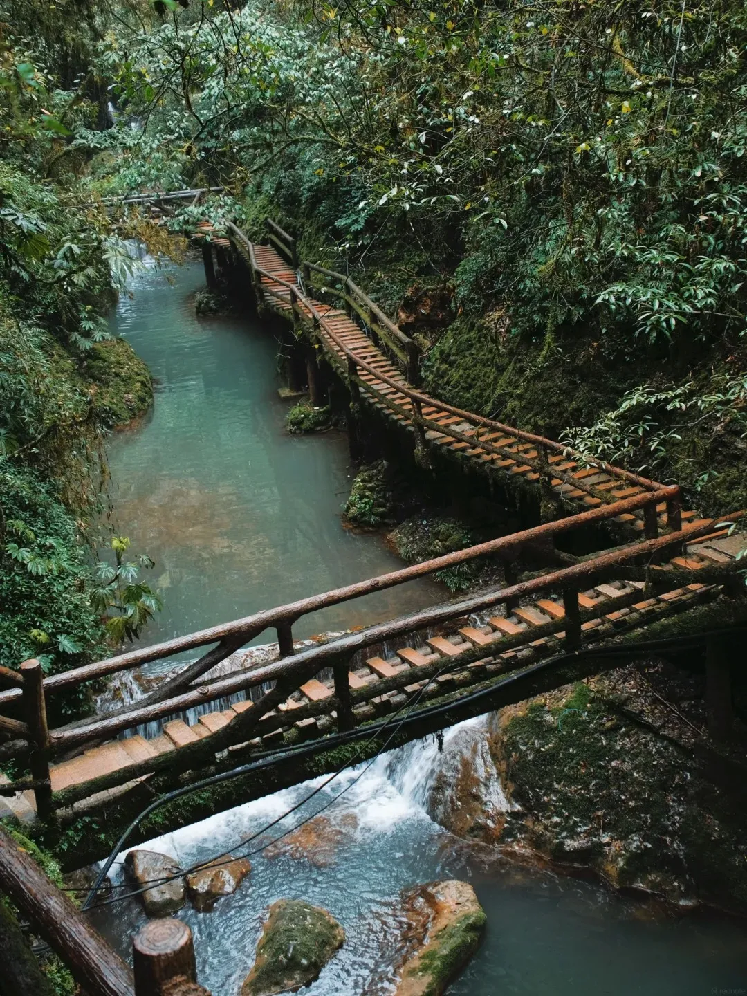 A wooden footbridge with a railing crosses a clear blue river flowing over rocks and small waterfalls, surrounded by lush green forest.