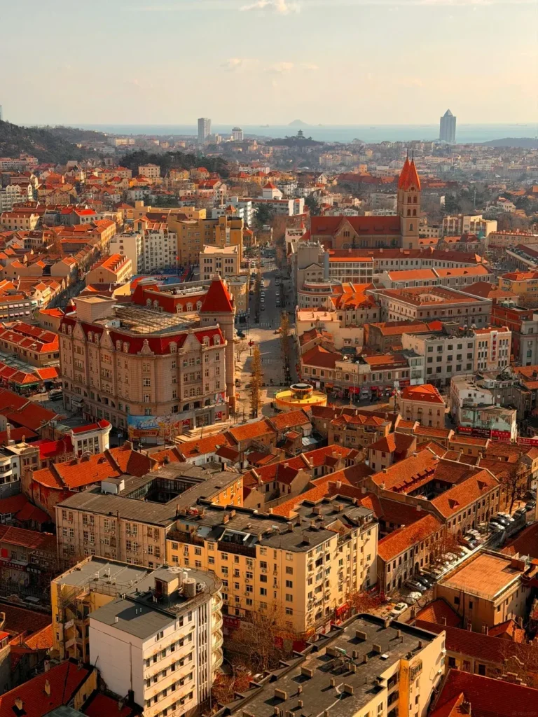 An aerial view of a densely packed city with numerous buildings featuring red tiled roofs. In the distance, a body of water, likely the sea, is visible under a bright sky, with several modern skyscrapers and a hill with a building on top dotting the horizon.