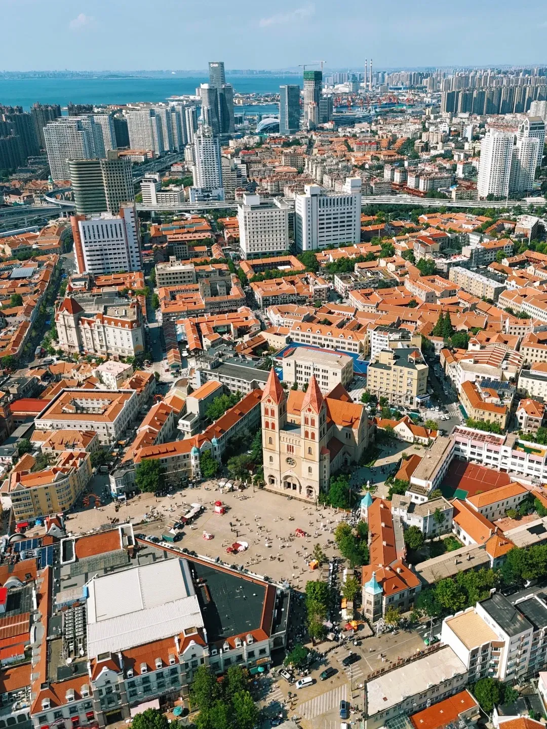 Aerial view of a large European-style cathedral with red-tiled roofs and twin towers, situated in a bustling public square in Qingdao. The square is surrounded by buildings with similar red-tiled roofs and European architectural influences. In the background, a modern cityscape with tall skyscrapers and a body of water can be seen.