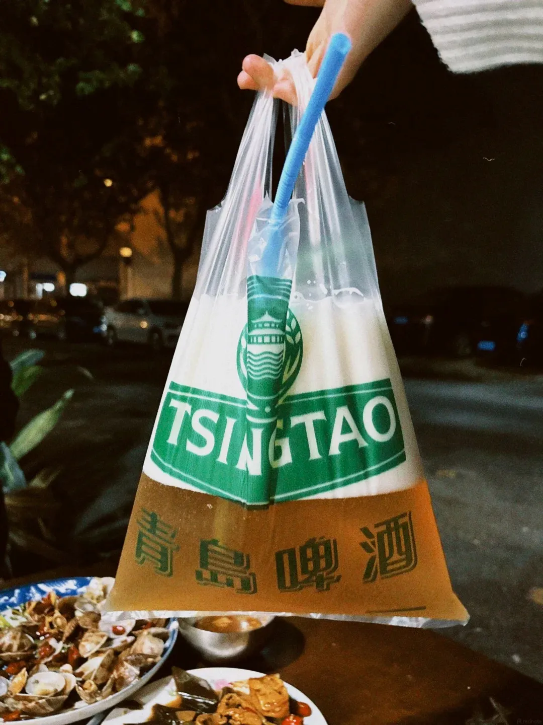 A person holds a clear plastic bag filled with Tsingtao beer, with a blue straw inserted. The bag is transparent, showing a lighter frothy liquid at the top and darker golden beer at the bottom. A green band with white text 'TSINGTAO' is visible on the bag, above Chinese characters. In the foreground, out of focus, are plates of food, including clams and other dishes, on a wooden table. The background is dark, suggesting an outdoor evening setting with trees and streetlights.