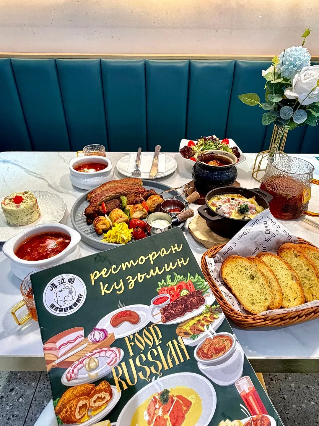 A table laden with a Russian-inspired meal, including a large platter of grilled meats, various bowls of soup and stew, a salad, bread basket with sliced garlic bread, and a menu featuring illustrations of Russian dishes.