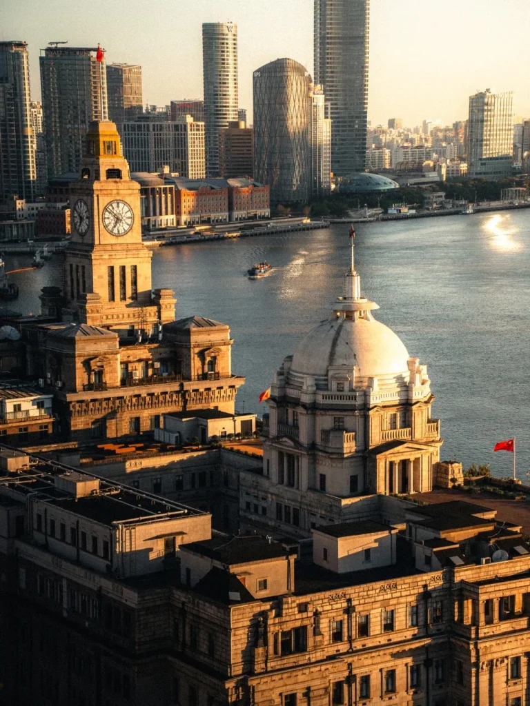 A panoramic aerial view of the Bund in Shanghai at sunset. In the foreground, the prominent clock tower of a historic colonial building stands tall, casting long shadows. Adjacent to it is a grand white dome structure. The Huangpu River reflects the golden light of the setting sun, with a small boat navigating its waters. In the background, a dense collection of modern skyscrapers pierces the sky, some illuminated by the sunset.