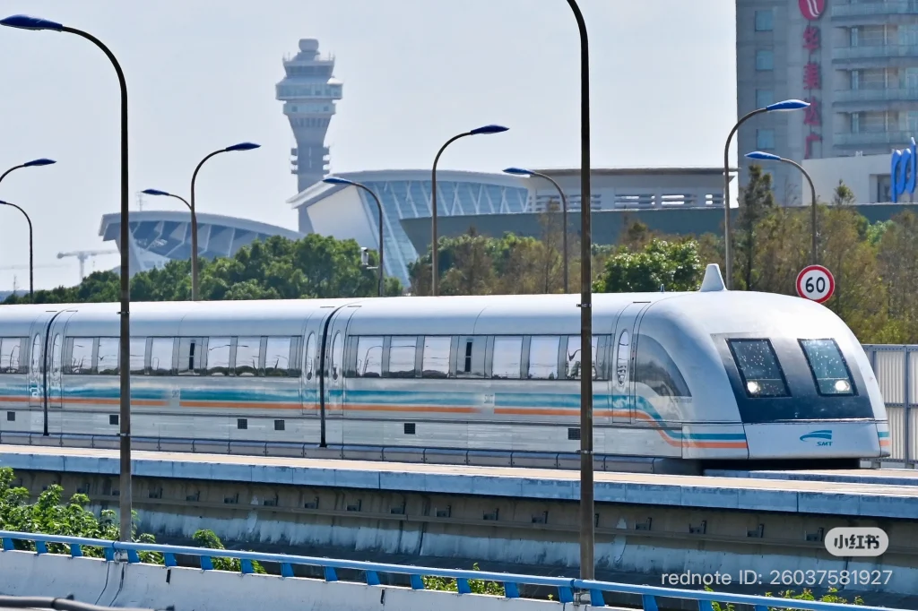 A silver and blue Shanghai Maglev train with orange and blue stripes approaching a station. Several tall streetlights with blue tops are visible in the foreground. In the background, an airport control tower and modern buildings can be seen under a clear sky.