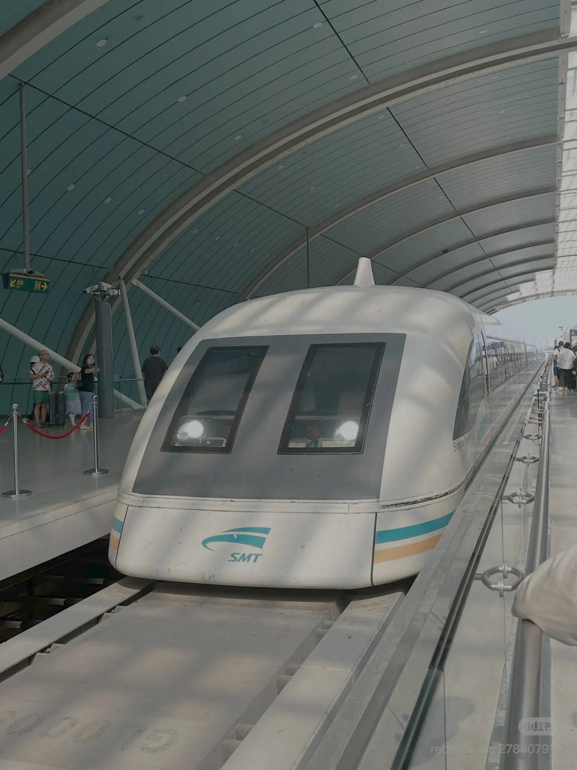 The front of a white Shanghai Maglev train with the SMT logo visible, arriving at a station platform. The train has large, dark-tinted windows and distinctive horizontal blue and orange stripes near the front. The station has a curved, teal-colored ceiling with white structural supports and modern lighting. Several people are visible on the platform, some waiting to board.