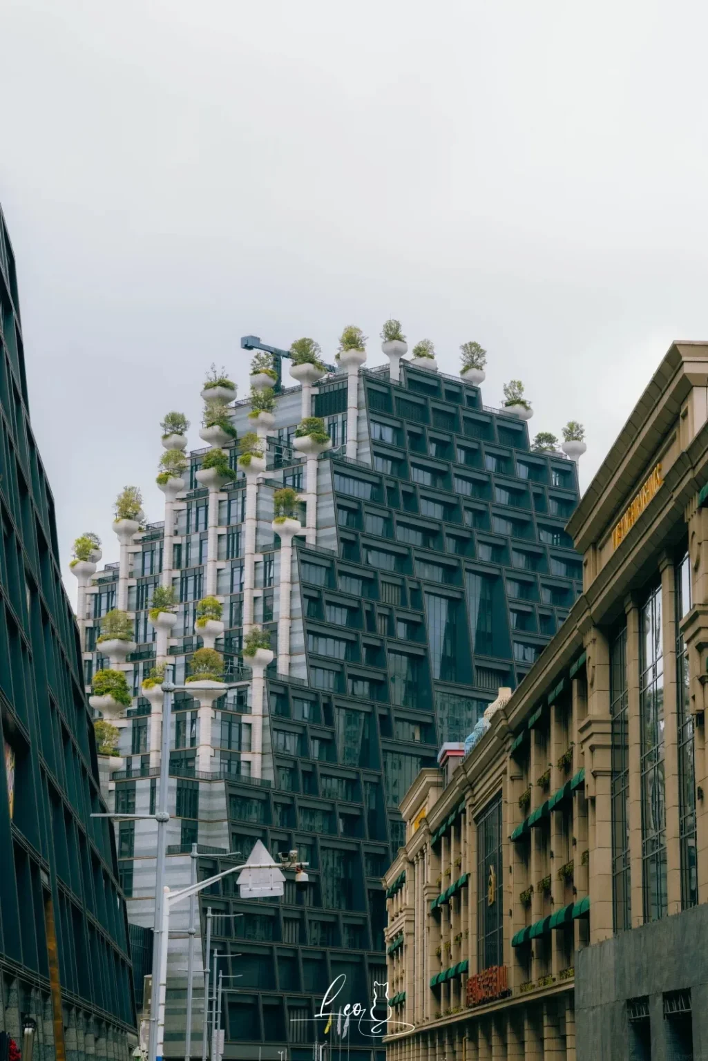 A unique modern skyscraper with a stepped facade is adorned with numerous white cylindrical planters, each holding a lush green tree. The building is surrounded by other urban structures, including a dark, multi-story building on the left and a classical-style building with columns and green awnings on the right. The sky above is overcast.