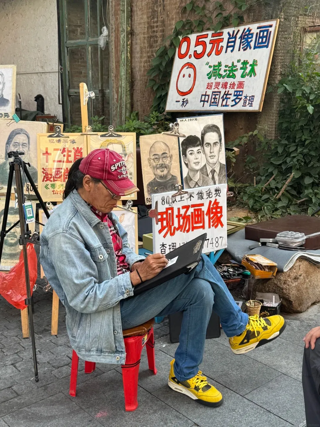 A person with a red baseball cap and denim jacket sits on a red stool, drawing in a black sketchbook. They are surrounded by various portraits and signs, including one advertising '0.5 Yuan Portrait' and another showing a couple. The artist is wearing bright yellow shoes and blue jeans.